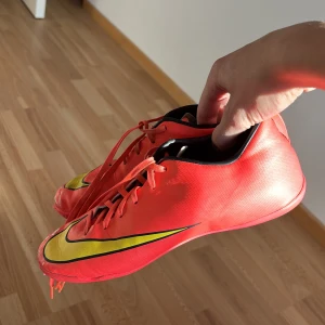Röda mercurials från Nike - Säljer ett par röda fotbollsskor från Nike med gula detaljer och snörning. Skorna har en slimmad passform och är perfekta för inomhusspel. Sulan är designad för optimalt grepp på inomhusplaner.
