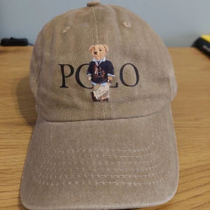 Beige keps från polo - Säljer en beige keps från Polo med en broderad björn på framsidan. Kepsen har justerbart spänne baktill och är tillverkad i bomull. Perfekt för en avslappnad stil.