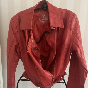 Röd skinn imitation jacka från O&S Paris - Snygg röd skinnjacka från O&S Paris med dragkedja och knappar framtill. Storlek S, det står L men det är italienska storlekar .Jackan har en stilren design med långa ärmar och quiltade detaljer på axlarna. Perfekt för att ge din outfit en cool touch.