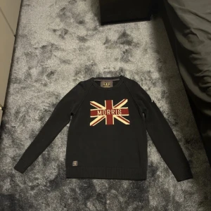 Svart tröja från Morris - Säljer en svart tröja från Morris med ett stilrent tryck av Union Jack och texten 'Morris' på bröstet. Tröjan är tillverkad i 100% bomull och har långa ärmar. Perfekt för en klassisk och avslappnad stil.