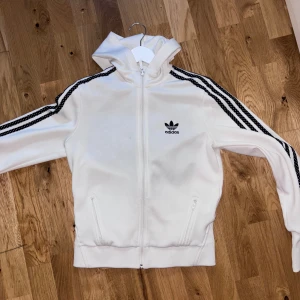 Vit hoodie från Adidas - Säljer en snygg vit hoodie från Adidas med dragkedja och klassiska svarta ränder längs ärmarna. Hoodien har en justerbar huva och två praktiska fickor framtill. Perfekt för en sportig look eller avslappnad stil.