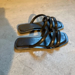 Svarta sandaler - Säljer ett par svarta sandaler med tre breda, vadderade remmar över foten och fyrkantig tå. Snygg minimalistisk design som passar till många outfits. Platt sula och öppen tå för en luftig känsla. Använda max 2 ggr som ni ser klistermärkena är kvar. Köpta i Danmark, Nypris 300kr.
