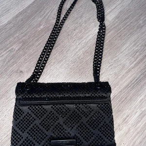 Svart axelväska från Kurt Geiger - Snygg svart axelväska från Kurt Geiger med kedjerem och coolt fågelhuvud som spänne. Väskan är täckt av svarta paljetter i rutmönster och har en lyxig satinliknande insida. Perfekt för dig som vill sticka ut med en edgy accessoar.