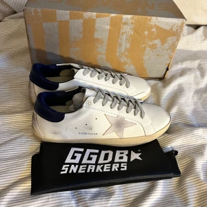 Golden Goose superstar  - Golden Goose sneakers i vitt skinn med grå mockastjärna på sidan och mörkblå häl. Grå snörning och beige sula med vintage-look. Klassisk låg modell med GGDB/SSTAR-tryck och originalpåse. Perfekta för en cool och avslappnad stil. Obs lnte 0rglnal 