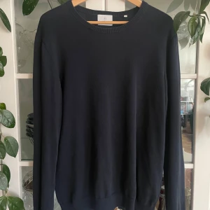 Mörkblå stickad tröja från Bläck - En stilren mörkblå stickad tröja från Bläck med rund halsringning och långa ärmar. Tröjan har en klassisk passform och ribbade muddar vid hals, ärmslut och nederkant. Perfekt för lager-på-lager och enkel att matcha med allt.