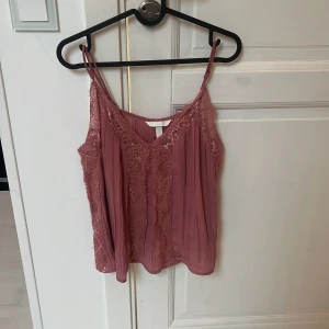 Rosa linnetopp med spets från H&M - Supersöt rosa linnetopp från H&M med tunna axelband och v-ringning. Toppen har fina spetsdetaljer längs kanterna och framtill, samt plisserat tyg som ger en luftig känsla. Perfekt för varma dagar eller när du vill känna dig extra fin.