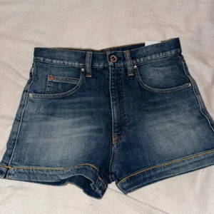 Armani Jeans denimshorts med strass - Snygga blå denimshorts från Armani Jeans med hög midja och klassisk femficksdesign. Baksidan har coola strassdetaljer i rött och silver på fickorna samt logga. Shortsen har gul kontrastsöm och är perfekta för sommaren. Riktigt trendiga och lätta att matcha.