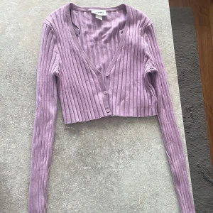 Ljuslila croppad kofta från MONKI - Superfin ljuslila kofta från MONKI med croppad passform. Tröjan är ribbad, har v-ringning och knäpps med tre knappar framtill. Långärmad och perfekt att styla över ett linne eller t-shirt för en chill look.