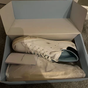 Philippe Model vita sneakers i skinn - Snygga vita Philippe Model sneakers i skinn med svarta detaljer på hälen och vintage-inspirerade slitningar. Klassisk låg modell med snörning, rund tå och platt sula. Perfekta för dig som gillar en clean men edgy look. De är dessutom helt oanvända. Frakt kommer att tillkomma 