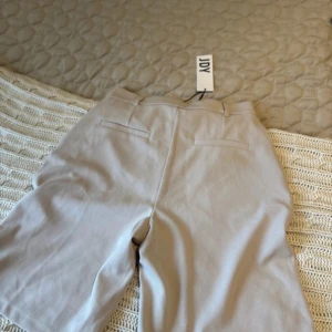 Beige bermudashorts från JDY - Stilrena beige bermudashorts från JDY med hög midja och klassisk passform. Shortsen har hällor för bälte, diskreta bakfickor och är tillverkade i ett mjukt bomullsmaterial. Perfekta för en clean och avslappnad look under varmare dagar.