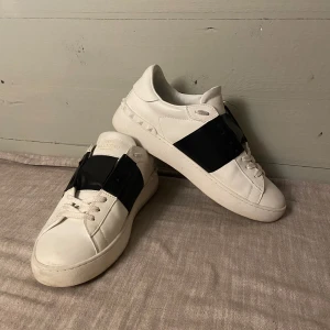 Valentino Garavani Open sneakers vit/svart - Snygga Valentino Garavani Open sneakers i vitt skinn med bred svart läderdetalj över vristen. Klassisk rund tå, snörning och ikoniska nitar baktill på sulan. Perfekta för dig som gillar stilrena och lyxiga sneakers med en twist.