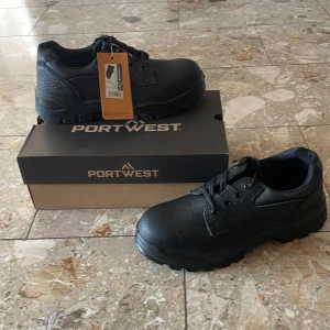 Svarta Portwest arbetsskor i skinn - Helt nya stabila svarta arbetsskor från Portwest med grov sula och snörning. Skorna är tillverkade i tåligt skinn och har en klassisk rund tå med stålhetta. Perfekta för dig som vill ha en robust och stilren look på jobbet eller i vardagen.