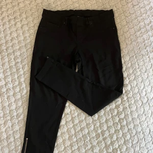 Svarta skinny byxor med dragkedja - Svarta byxor i skinny modell med dragkedjedetalj vid benslut. Klassisk femficksdesign och bälteshällor. Snygg passform som sitter tight hela vägen ner. Perfekta till sneakers eller boots.