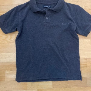 Mörkgrå pikétröja från Bondelid - Snygg mörkgrå pikétröja från Bondelid med klassisk krage och två knappar framtill. Tröjan har korta ärmar och diskret broderad logga på bröstet. Perfekt för en clean och stilren look.