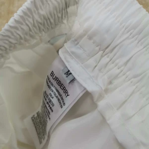 Vita Burberry shorts med rutiga sidor - Snygga vita shorts från Burberry med klassiska rutiga paneler på sidorna och guldfärgat broderi framtill. De har elastisk midja med snörning för perfekt passform. Perfekta för sommaren och ger en lyxig känsla till din outfit.