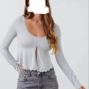 Ljusgrå croppad topp med spetskant - Superfin ljusgrå croppad topp med lång ärm och söt spetskant längs ringningen. Toppen har en liten v-ringning och en liten rosett framtill som ger en öppen detalj. Nästan aldrig använd 