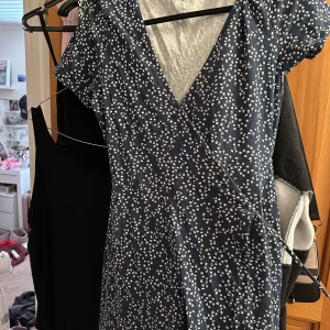 Blå omlottklänning med vita blommor - Supersöt kort omlottklänning i marinblått med små vita blommor. Klänningen har korta ärmar, v-ringning och knytband i midjan för en snygg passform. Perfekt för varma dagar och har en lätt, flowy känsla.