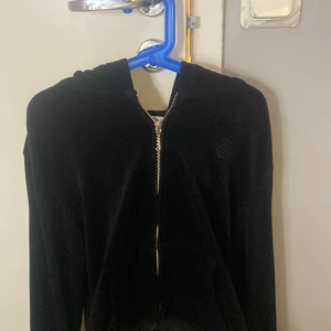Svart velourjacka Juicy Couture - Svart velourjacka från Juicy Couture med dragkedja framtill och huva. Klassisk broderad logga på bröstet och mjukt material som ger en lyxig känsla. Perfekt för en chill och trendig look.