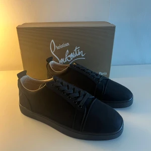 Svarta sneakers från Christian Louboutin - Det här är ett par lyxiga sneakers från märket Christian Louboutin. De är svarta med en mjuk insida och har märket skrivet i guld på sulan inuti. Undersidan är röd, vilket är Louboutins kända stil. Skorna passar bra till både vardag och finare tillfällen.