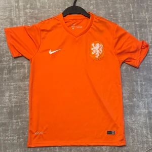 Holländsk Nike fotbollströja orange - Snygg orange fotbollströja från Nike med Nederländernas lejonemblem på bröstet och vit Nike-logga. Tröjan är tillverkad i lätt och ventilerande Dri-FIT-material och har V-ringning samt korta ärmar. Perfekt för dig som älskar fotboll och vill sticka ut på planen.
