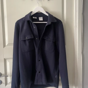 Mörkblå overshirt från Selected Homme - Stilren mörkblå overshirt från Selected Homme med klassisk krage, två stora bröstfickor med lock och tryckknappar. Skjortan har långa ärmar och en rak passform, perfekt att bära som lager. Materialet känns mjukt och är troligtvis bomull eller polyester.