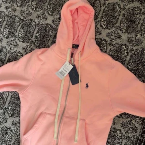 Ljusrosa hoodie från Polo Ralph Lauren - Superfräsch ljusrosa hoodie från Polo Ralph Lauren med dragkedja och klassisk logga broderad på bröstet. Tröjan har huva med snörning, känguruficka och långa ärmar. Perfekt för en chill och snygg look.