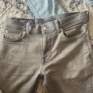 Grå stretchjeans Tommy Hilfiger  - En hel del utdragna efter mycket användning i midjan! Du ser på bild 3 vid fickan lite slitning men de är all slitning jeansen har.                Snygga grå jeans från Tommy Hilfiger med TH Flex advanced stretch för extra komfort. Klassisk femficksmodell med diskreta röda detaljer och logga på bakfickan. Perfekta för dig som vill ha stilrena och bekväma jeans med modern passform.