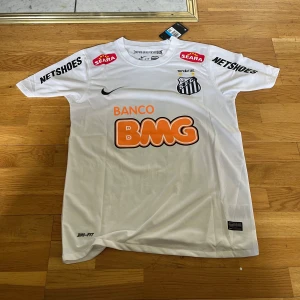 Vit Santos Neymar Jr fotbollströja Nike - Santos hemmatröja med Neymar Jr och nummer 11 på ryggen. Vit med svarta och orange tryck, flera sponsorer och klubbmärke på bröstet. Tillverkad i lätt Dri-FIT-material som andas. Klassisk passform och korta ärmar, perfekt för fotbollsälskare.
