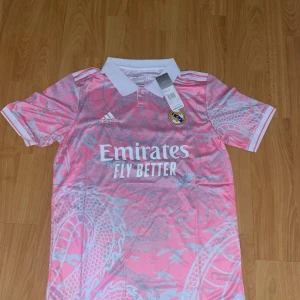 Rosa Real Madrid fotbollströja Adidas - Snygg rosa Real Madrid fotbollströja från Adidas med vit krage och detaljer. Tröjan har korta ärmar och ett unikt drakmönster i ljusgrått. Klubbmärke och Adidas-logga på bröstet samt Emirates Fly Better-tryck på framsidan.