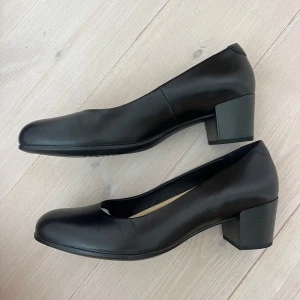 Svarta pumps från ECCO i skinn - Stilrena svarta pumps från ECCO i mjukt skinn med rund tå och låg, stabil klack. Klassisk design med enkel siluett och diskret ECCO-logga på innersulan.