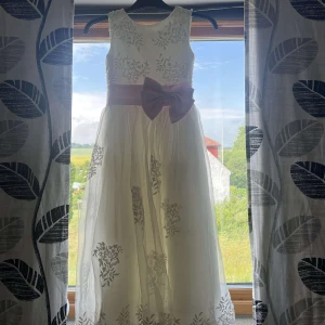Vit långklänning med rosa rosett - Superfin vit långklänning med broderade blommor och glittriga detaljer. Klänningen har en bred ljusrosa satinrosett i midjan och är ärmlös. Kjolen är i flera lager med transparent tyg och vackra broderier längst ner. Perfekt för speciella tillfällen! Endast använd 1 gång, till barn i 5-7 års åldern! är lite smutsig längst ner men inget som syns