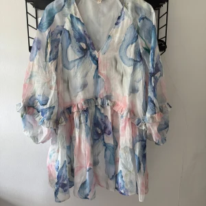 Blommig kortklänning med volang - Superfin kortklänning med blommigt mönster i pastelliga toner av blått, rosa och vitt. Klänningen har v-ringning, volangdetaljer och vida, långa ärmar. Tunt, luftigt material som känns perfekt för varmare dagar.