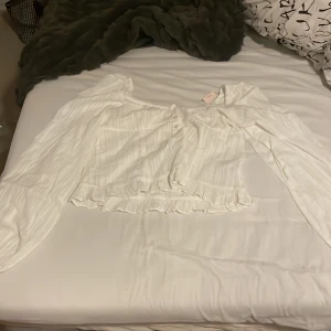 Vit blus med puffärmar från Nelly - Supersöt vit blus från Nelly med långa ärmar och offshoulder-modell. Blusen har knappar framtill, volangkant nertill och ett lätt ribbat tyg med diskret randigt mönster. Perfekt för dig som gillar romantisk och trendig stil. HELT NY ALDRIG ANVÄND