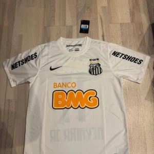 Vit Santos Neymar Jr fotbollströja Nike - Santos FC fotbollströja i vitt med Neymar Jr och nummer 11 på ryggen. Tröjan har korta ärmar, svarta tryck och orange BMG-logga. Tillverkad i lätt och ventilerande material, perfekt för match eller träning. Klassisk Nike Swoosh på bröstet och klubbmärke.