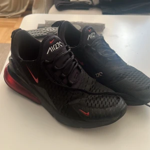 Nike Air Max 270 svarta sneakers - Svarta Nike Air Max 270 sneakers med röda detaljer och stor synlig Air-enhet i hälen. Ovandelen är i mesh för bra andningsförmåga och har snörning framtill. Snygg sportig look med vita och röda loggor på plösen och sidorna. Perfekta för dig som gillar streetstyle.