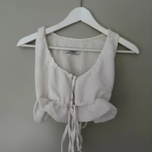 Vit linnetopp med snörning - Superfin vit linnetopp med snygg snörning framtill och volangkant nedtill. Toppen är ärmlös och har en lätt strukturerad yta som ger en cool känsla. Perfekt för varma dagar och enkel att styla med jeans eller kjol.