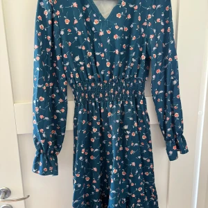 Blommig blå långärmad klänning - Supersöt blå klänning med små blommor i rött och vitt. Klänningen har v-ringning, långärmade ärmar med resår vid handlederna och smock i midjan för en snygg passform. 