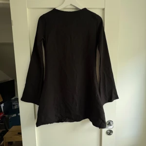 Svart kortklänning med öppen rygg - Svart kortklänning i ett mjukt tyg med långa, lätt utsvängda ärmar och en djup öppen rygg som knyts med band i nacken. Enkel och stilren design som ger en cool vibe. Perfekt för dig som gillar minimalistiskt men ändå vill sticka ut.