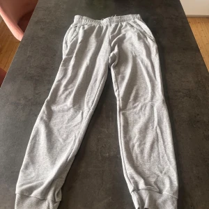 Grå mjukisbyxor från Adidas - Gråa mjukisbyxor från Adidas med resår i midjan och muddar vid bensluten. Byxorna har två sidofickor och är tillverkade i mjuk bomullsmix. Perfekta för chill eller träning, klassisk och enkel stil.