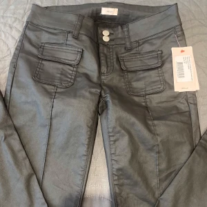 Low waist bootcut pocket jeans  - Low waist bootcut pocket jeans och två stora fickor framtill. Byxorna är lågmidjade, dubbla knappar och är tillverkade i ett glansigt material som ger en cool look. Perfekta för dig som vill ha en trendig och edgy stil.
