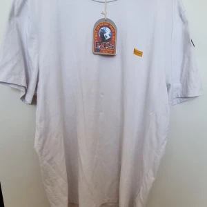 Vit iconic t-shirt från Parajumpers - Vit t-shirt från Parajumpers med klassisk passform. T-shirten har korta ärmar, ett gult broderat märke på bröstet och en stor Parajumpers-patch på ärmen. Tillverkad i mjuk bomull för en skön känsla.