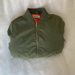 Olivgrön bomberjacka från H&M - Snygg olivgrön bomberjacka från H&M med orange foder. Jackan har dragkedja framtill, två fickor med dragkedja och en ärmficka. Klassisk ribbad krage, muddar och nederkant. Perfekt för dig som vill ha en stilren och trendig look.