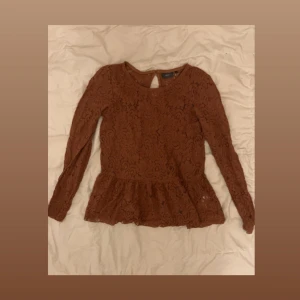 Brun spetsblus med volang - Säljer en brun långärmad blus i spets med snygg volang nertill. Blusen har rund halsringning och ett litet knappslå bak i nacken. Perfekt för dig som gillar romantiska detaljer och vill sticka ut lite extra.