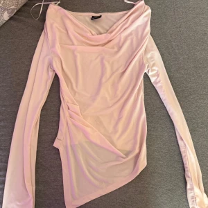 Ljusrosa omlottopp från Glamorous - Supersnygg ljusrosa omlottopp från Glamorous med draperad halsringning och lång ärm. Toppens asymmetriska nederkant och tunna, stretchiga material ger en modern vibe. Perfekt att styla offshoulder för en cool look.