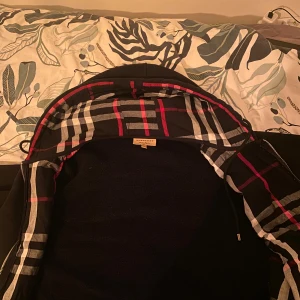 Svart Burberry hoodiejacka med rutor - Svart hoodiejacka från Burberry med klassiskt rutigt foder i huvan i svart, vitt och rött. Jackan har dragkedja framtill och långa ärmar. , perfekt för dig som vill ha en stilren och ikonisk look. Säljer denna för har gymmat och blivit lite för stor för denna. Mycket bra skick 8/10/// använd 2 gånger tvättat en gång. Pris kan diskuteras 