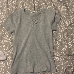 Grå ribbad topp med snörning - Grå ribbad topp från Gina Tricot med korta ärmar och snygg snörningsdetalj vid halsringningen. Modellen är figurnära och har en mjuk, stretchig känsla som gör den superbekväm. Perfekt att styla med jeans eller kjol för en chill look.