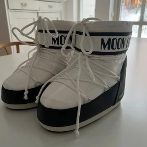 Svarta och vita Moon Boots - Moon Boots i svart och vitt med klassisk logga runt skaftet. Skorna har snörning framtill och en rejäl platt sula. Perfekta för vintern med sin vadderade och syntetiska ovandel som håller fötterna varma. Rymdinspirerad design som sticker ut.