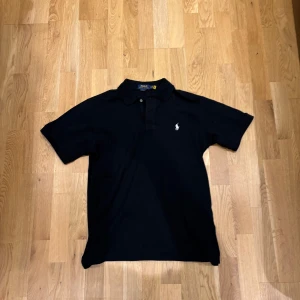 Svart pikétröja från Ralph Lauren - Klassisk svart pikétröja från Ralph Lauren med vit broderad logga på bröstet. Tröjan har korta ärmar, krage och knäppning framtill. Perfekt basic-plagg med snygg passform och tidlös stil. Ny skick! Nypris runt 1000