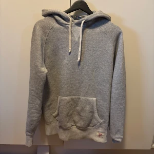 Grå hoodie från Polo Ralph Lauren - Klassisk grå hoodie från Polo Ralph Lauren med huva och snörning, stor känguruficka framtill och diskret logolapp vid nederkanten. Mjuk insida och relaxed fit, perfekt för chill dagar. Tillverkad i bomullsblandning för extra komfort.