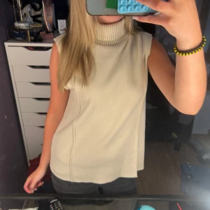 Beige  polotopp  - Säljer en beige ärmlös polotopp i stickat material. Toppen har hög polokrage och en rak, lite loose passform. Perfekt att styla med jeans eller kjol för en clean och chill look.